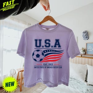 May include: A light purple short-sleeve t-shirt featuring a graphic with the text U.S.A. above a soccer ball and a stylized red, white, and blue flag design. Below the graphic, the text reads Est. 1913 United States of America Soccer Club.
