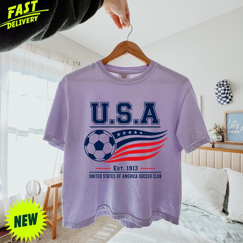 May include: A light purple short-sleeve t-shirt featuring a graphic with the text U.S.A. above a soccer ball and a stylized red, white, and blue flag design. Below the graphic, the text reads Est. 1913 United States of America Soccer Club.