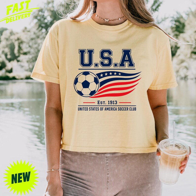 May include: A pale yellow short-sleeved t-shirt featuring a graphic with the text U.S.A. above a soccer ball and stylized red and blue flag stripes. Below the graphic, the text reads EST. 1913 UNITED STATES OF AMERICA SOCCER CLUB.