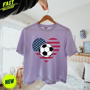 Puede incluir: Una camiseta de manga corta de color morado claro con un gr&aacute;fico central en forma de coraz&oacute;n con el dise&ntilde;o de la bandera estadounidense y un bal&oacute;n de f&uacute;tbol en blanco y negro en el centro. La prenda tiene cuello redondo y corte holgado.