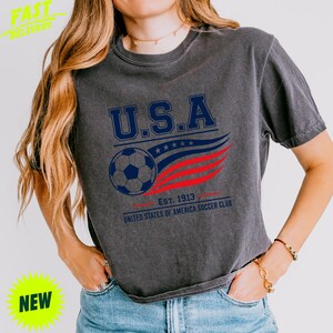 May include: A charcoal grey short-sleeve t-shirt featuring a graphic with the text U.S.A. above a soccer ball and red and blue wavy lines. Below the graphic, the text reads EST. 1913 UNITED STATES OF AMERICA SOCCER CLUB in dark blue lettering.