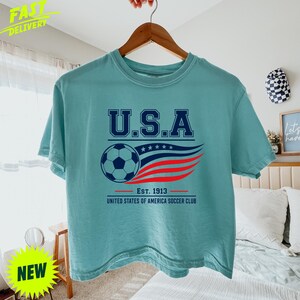 May include: A light teal short-sleeve t-shirt featuring a graphic with the text U.S.A. above a soccer ball and stylized red and blue wavy lines. Below the graphic, the text reads EST. 1913 UNITED STATES OF AMERICA SOCCER CLUB in dark blue.