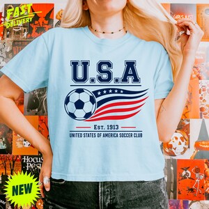 May include: A light blue short-sleeve t-shirt featuring a graphic print with the text U.S.A. above a soccer ball and stylized red and blue stripes. Below the graphic, the text reads EST. 1913 UNITED STATES OF AMERICA SOCCER CLUB.