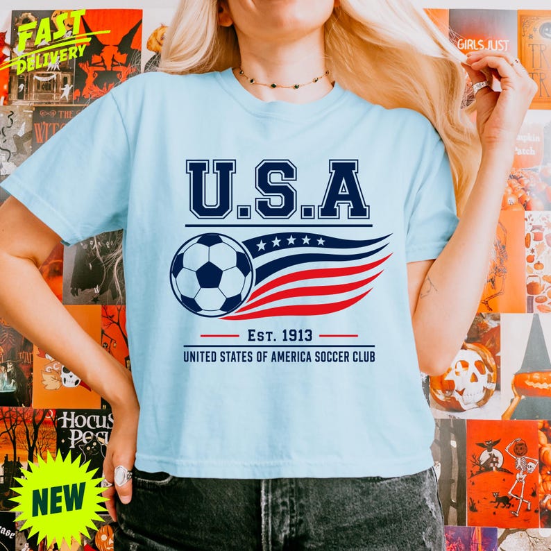 May include: A light blue short-sleeve t-shirt featuring a graphic print with the text U.S.A. above a soccer ball and stylized red and blue stripes. Below the graphic, the text reads EST. 1913 UNITED STATES OF AMERICA SOCCER CLUB.