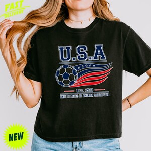 May include: A black short-sleeve t-shirt featuring a graphic with the text U.S.A. above a soccer ball and stylized red, white, and blue wavy lines. Below the graphic, the text reads EST. 1913 and UNITED STATES OF AMERICA SOCCER CLUB.