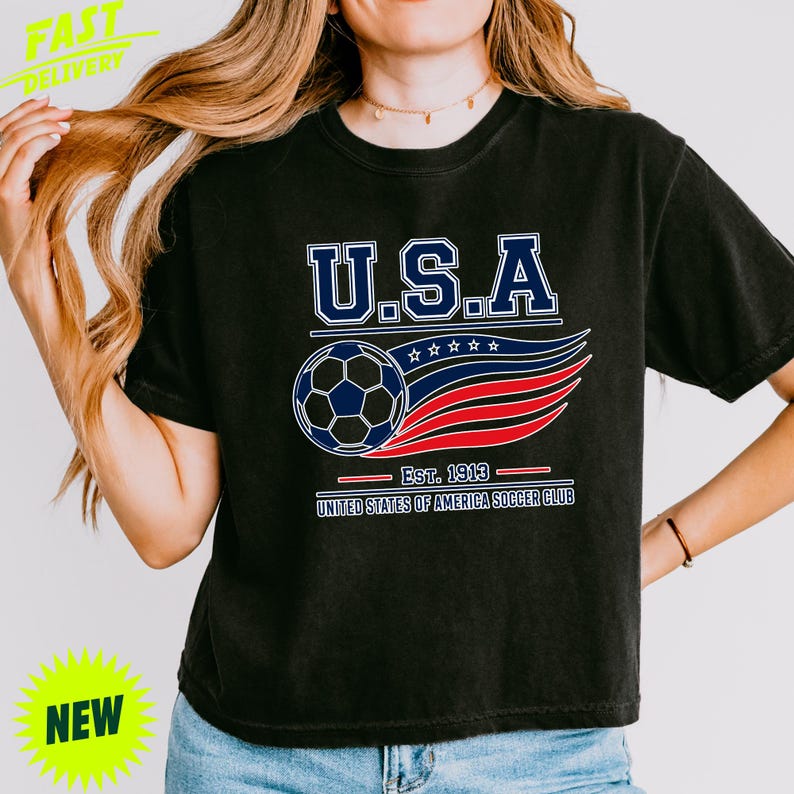 May include: A black short-sleeve t-shirt featuring a graphic with the text U.S.A. above a soccer ball and stylized red, white, and blue wavy lines. Below the graphic, the text reads EST. 1913 and UNITED STATES OF AMERICA SOCCER CLUB.