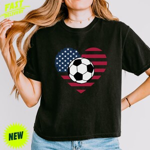 Puede incluir: Una camiseta negra de manga corta con un gr&aacute;fico en forma de coraz&oacute;n con los colores de la bandera estadounidense y un bal&oacute;n de f&uacute;tbol en el centro. El dise&ntilde;o incluye el campo azul con estrellas blancas a la izquierda y rayas rojas y blancas a la derecha.