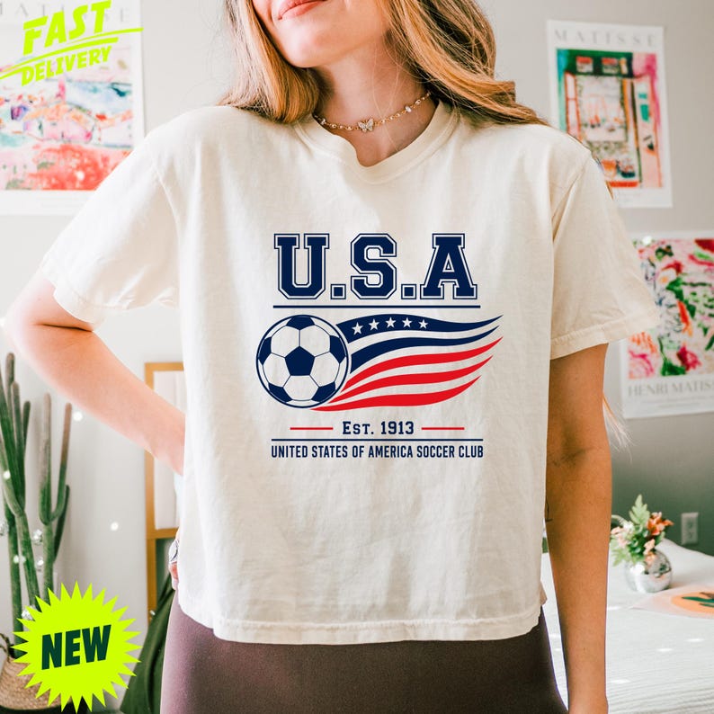May include: A cream-colored short-sleeve t-shirt featuring a graphic with the text U.S.A. above a soccer ball and a stylized red, white, and blue wave design. Below the graphic, the text reads EST. 1913 UNITED STATES OF AMERICA SOCCER CLUB.