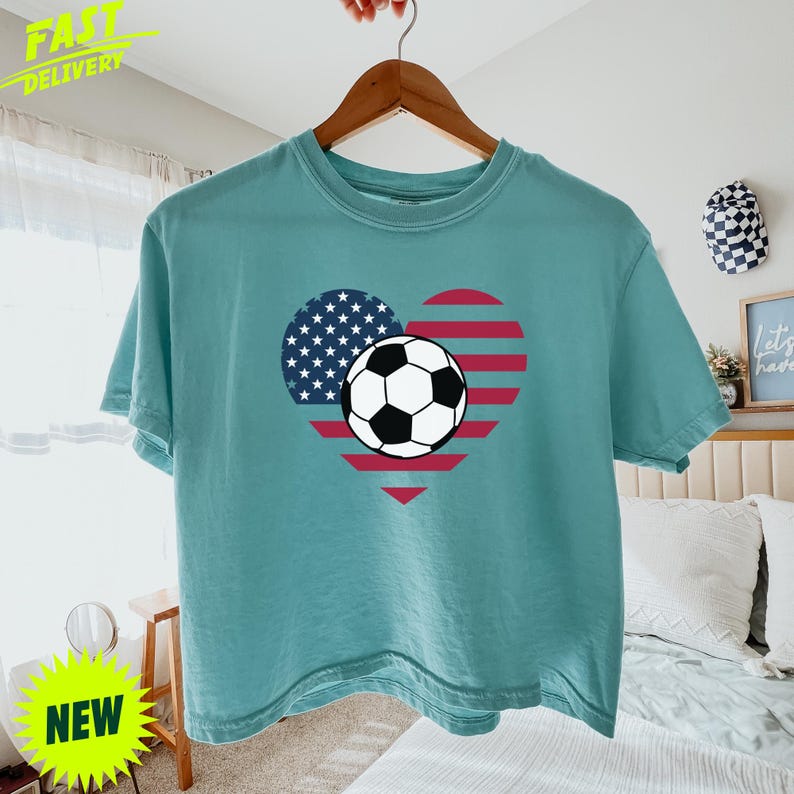 Puede incluir: Una camiseta de manga corta color verde azulado con un gr&aacute;fico de coraz&oacute;n relleno con el patr&oacute;n de la bandera estadounidense, incluyendo rayas rojas y blancas y una secci&oacute;n azul con estrellas blancas, centrado con un bal&oacute;n de f&uacute;tbol.