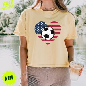 Puede incluir: Una camiseta de manga corta de color amarillo p&aacute;lido con un gr&aacute;fico de un coraz&oacute;n relleno con el patr&oacute;n de la bandera estadounidense y un bal&oacute;n de f&uacute;tbol en blanco y negro centrado sobre el dise&ntilde;o del coraz&oacute;n. Tiene cuello redondo.