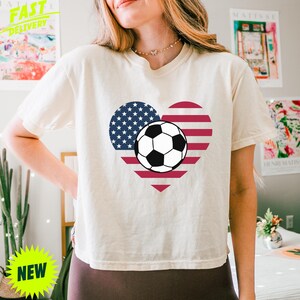 Puede incluir: Una camiseta de manga corta de color crema con un gr&aacute;fico central en forma de coraz&oacute;n relleno con el patr&oacute;n de la bandera estadounidense de rayas rojas y blancas y estrellas azules, con un bal&oacute;n de f&uacute;tbol blanco y negro en el centro.