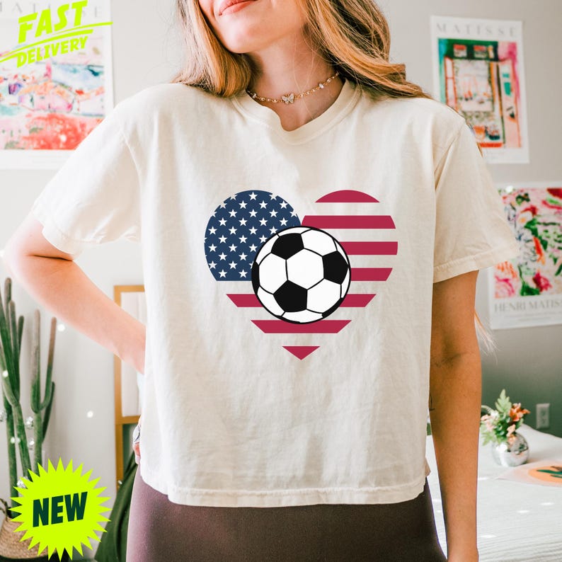 Puede incluir: Una camiseta de manga corta de color crema con un gr&aacute;fico central en forma de coraz&oacute;n relleno con el patr&oacute;n de la bandera estadounidense de rayas rojas y blancas y estrellas azules, con un bal&oacute;n de f&uacute;tbol blanco y negro en el centro.