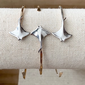 May include: Three silver-colored ray-shaped pendants on gold-colored curved bars. The pendants are displayed on a beige fabric surface. The jewelry is designed to be worn as earrings or a bracelet.