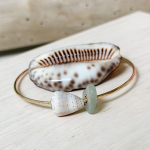 May include: A gold-toned metal bangle bracelet featuring a small, conical, speckled white shell bead and a smooth, pale sea-glass disc. The bracelet is positioned in front of a larger, oval-shaped cowrie shell with brown spots and a dark brown central slit.