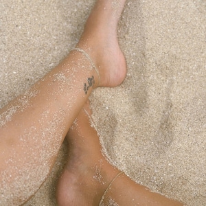 May include: Close-up of legs and feet covered in sand, with a delicate gold ankle bracelet. A small tattoo is visible on the ankle. The image evokes a beach or vacation theme, with a focus on jewelry and body art.