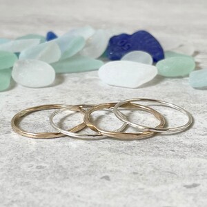 May include: Four simple stacking rings, two gold and two silver, with a hammered texture. The rings are stacked on a light gray surface.