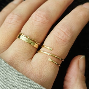 May include: Two gold rings on a finger. The bottom ring is a simple band, while the top ring is a textured band with a wavy edge.