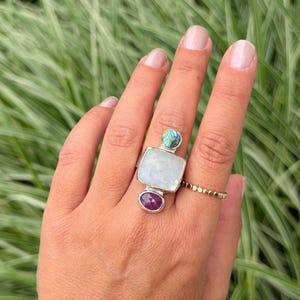 May include: A silver ring featuring a large square moonstone centre, a small round abalone shell accent at the top, and a small oval ruby at the bottom. A thin gold beaded band is worn on the adjacent finger, complementing the multi-stone design.