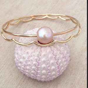 May include: A gold-colored bangle bracelet with a scalloped design, featuring a large, round, pink pearl. The bracelet rests on a light purple sea urchin. The text "jack & ginger" is visible on the bracelet.