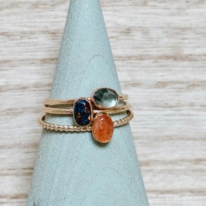 May include: Three gold rings with different colored gemstones. The top ring has a light blue gemstone, the middle ring has a dark blue and black gemstone, and the bottom ring has a light orange gemstone.