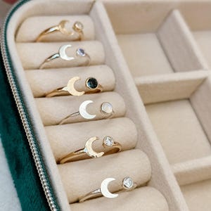 May include: A close-up of a green velvet jewelry box with six compartments. Each compartment holds a silver or gold ring with a crescent moon design and a small gemstone.