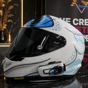 Könnte beinhalten: Ein weißer Integral-Motorradhelm mit blauen und hellblauen Grafikmustern, einem dunkel getönten Visier und einem schwarzen Kommunikationsgerät an der Seite. Die Helmschale zeigt ein stilisiertes Augendesign im oberen und seitlichen Bereich.