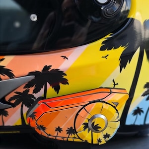 Könnte beinhalten: Ein Motorradhelm mit einem lebendigen tropischen Sonnenuntergangsmotiv, schwarzen Palmensilhouetten, orangefarbenen und gelben Diagonalstreifen sowie kleinen Vögeln. Ein passendes Kommunikationsgerät ist an der Seite angebracht und zeigt dasselbe Muster.