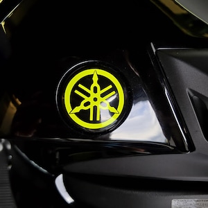 Könnte beinhalten: Nahaufnahme eines schwarzen Motorradteils mit einem kreisförmigen Yamaha-Emblem. Das Logo ist ein gelbes, stilisiertes Design in einem schwarzen Kreis. Die Karosserie des Motorrads ist schwarz mit glänzenden Oberflächen.
