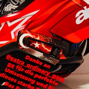 May include: A bright red motorcycle helmet featuring a white Alpinestars logo and a custom visor attachment. The attachment displays a red and white Turkish flag design with lightning patterns and the text AMO. Red German text overlays the image.