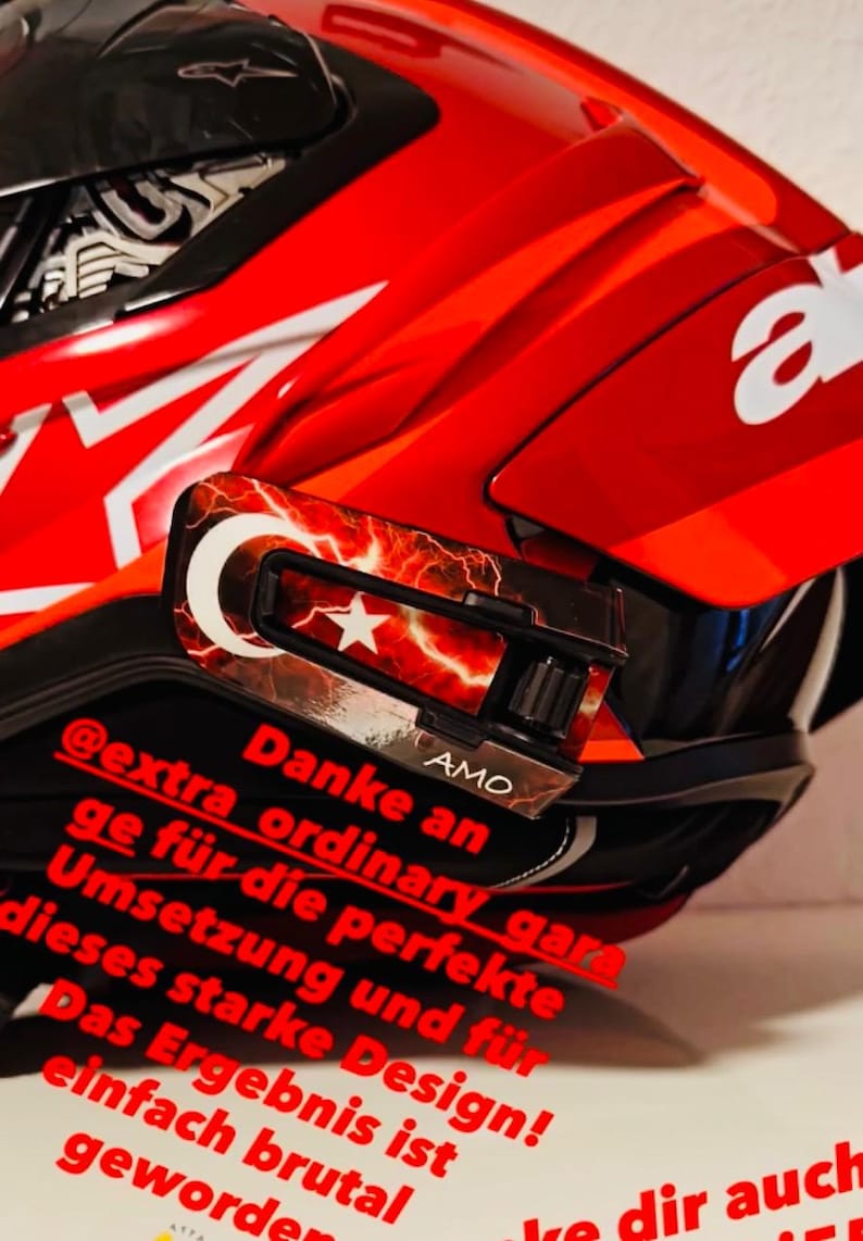 May include: A bright red motorcycle helmet featuring a white Alpinestars logo and a custom visor attachment. The attachment displays a red and white Turkish flag design with lightning patterns and the text AMO. Red German text overlays the image.