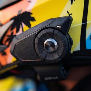 Może przedstawiać: Czarne urządzenie komunikacyjne Sena do kasku motocyklowego o teksturowanym wykończeniu, z centralnym okrągłym pokrętłem i srebrnym metalowym przyciskiem, zamontowane na kasku z ż&oacute;łto-pomarańczowo-czarnym wzorem graficznym w palmy.