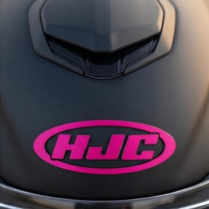 Może przedstawiać: Zbliżenie na matowy czarny kask motocyklowy z jasnoróżowym owalnym logo z napisem "HJC". Kask ma elegancką, aerodynamiczną konstrukcję z otworem wentylacyjnym na górze. Logo jest wyśrodkowane z przodu kasku.