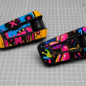 May include: Two motorcycle helmet communication system units featuring vibrant graphic skins. The left unit displays a blue and yellow monster face with pink X eyes. The right unit features a black background with colorful pink and blue paint drips.