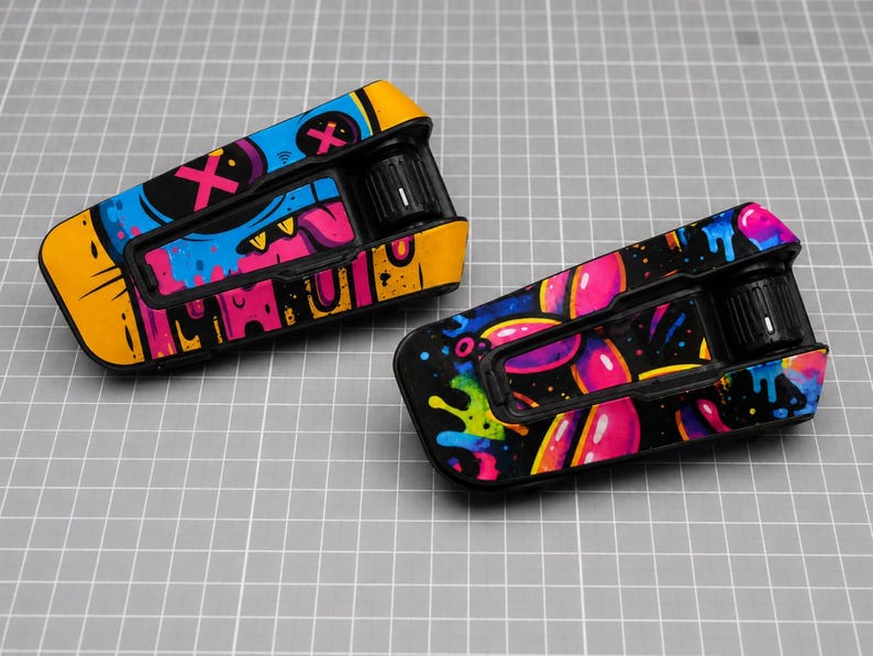 May include: Two motorcycle helmet communication system units featuring vibrant graphic skins. The left unit displays a blue and yellow monster face with pink X eyes. The right unit features a black background with colorful pink and blue paint drips.