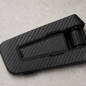 May include: A black phone stand featuring a carbon fiber pattern on its surface. The stand has a rectangular base with a hinged support arm and a cylindrical adjustment knob at the top, designed to hold a mobile device at an angle.