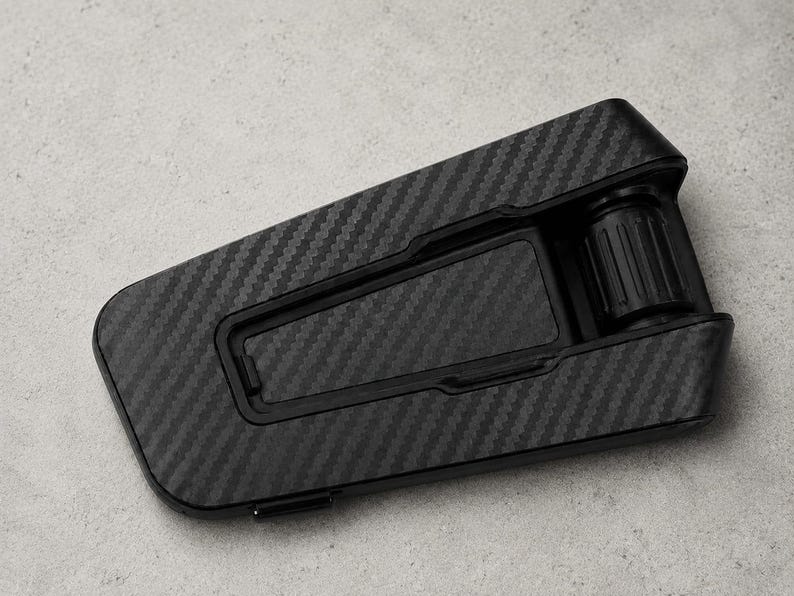 May include: A black phone stand featuring a carbon fiber pattern on its surface. The stand has a rectangular base with a hinged support arm and a cylindrical adjustment knob at the top, designed to hold a mobile device at an angle.