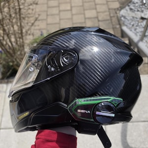 Może przedstawiać: Czarny kask motocyklowy z wł&oacute;kna węglowego z przezroczystym wizjerem oraz zielono-czarnym urządzeniem komunikacyjnym Sena przymocowanym z boku. Kask ma błyszczące wykończenie z widocznym splotem oraz pasek pod brodę z klamrą.