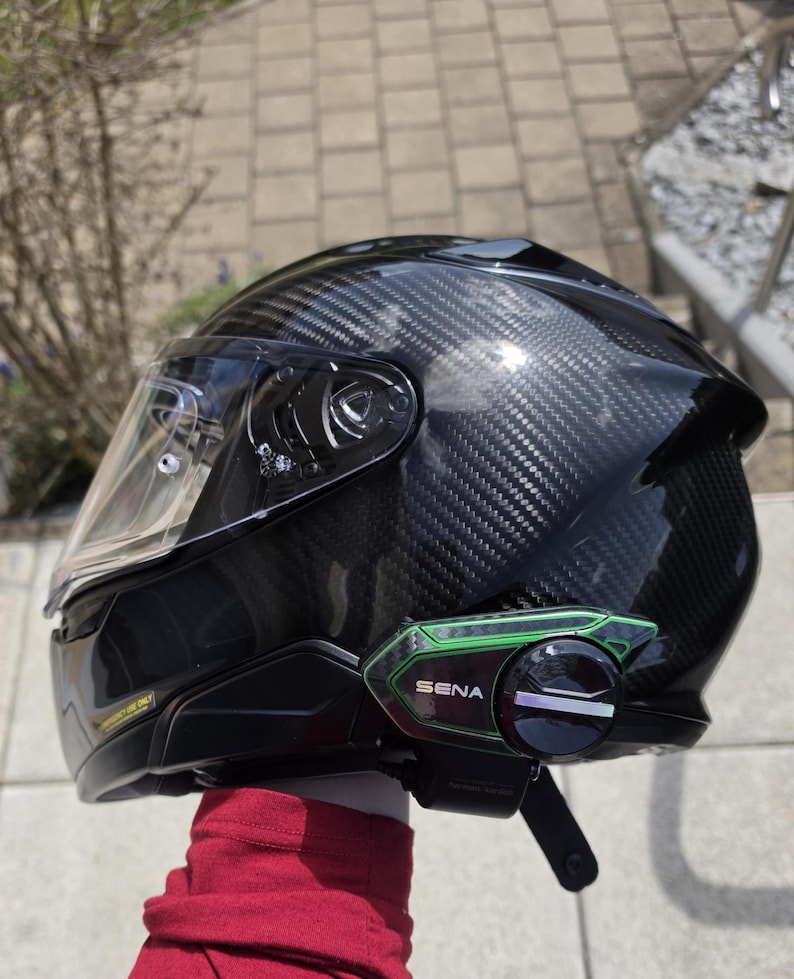 Może przedstawiać: Czarny kask motocyklowy z wł&oacute;kna węglowego z przezroczystym wizjerem oraz zielono-czarnym urządzeniem komunikacyjnym Sena przymocowanym z boku. Kask ma błyszczące wykończenie z widocznym splotem oraz pasek pod brodę z klamrą.