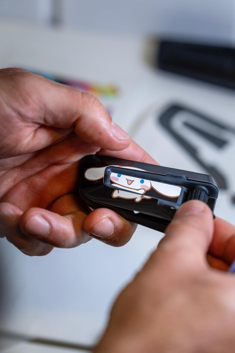 May include: A black plastic utility knife featuring a graphic of a white cartoon character with long ears, blue eyes, and a pink mouth. The tool has a sliding mechanism on the top and a textured black wheel on the side for blade adjustment.