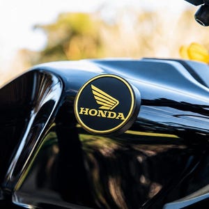 May include: A glossy black motorcycle fuel tank featuring a circular emblem with a gold-coloured Honda wing logo and the word HONDA printed in gold lettering. The emblem has a thin gold border and is mounted on the side of the curved black surface.