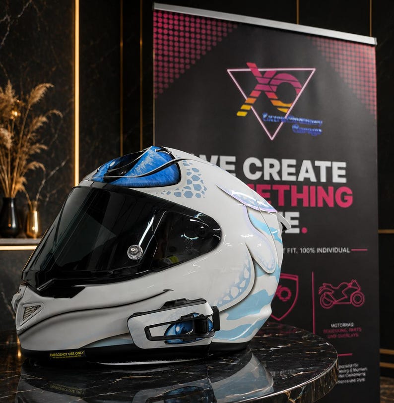 May include: A white full-face motorcycle helmet featuring blue and light blue graphic patterns, a dark tinted visor, and a black communication device mounted on the side. The helmet shell displays a stylized eye design near the top and side.