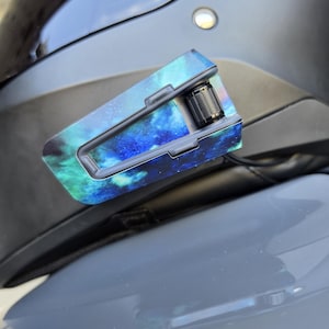 May include: A motorcycle helmet communication device attached to the side of a matte black helmet. The device features a vibrant blue and green nebula-patterned skin with a prominent black circular control dial on the rear section of the unit.