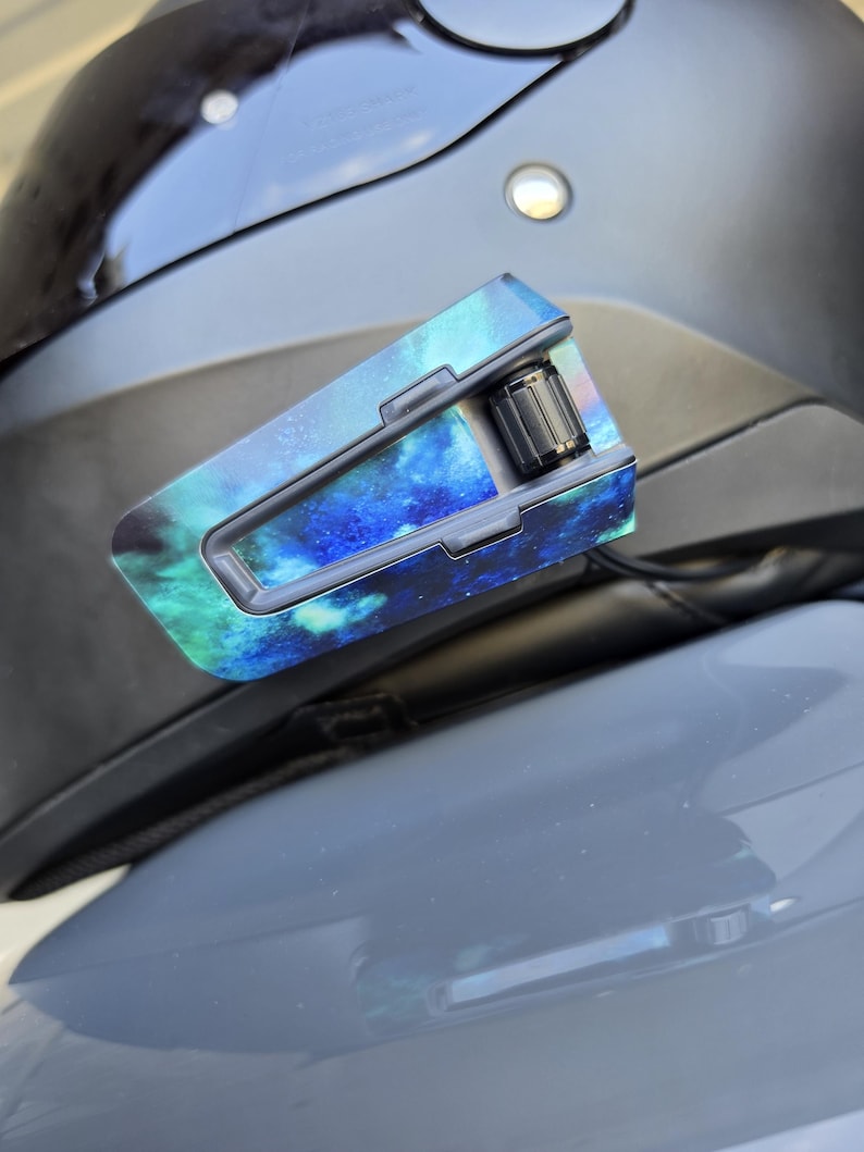 May include: A motorcycle helmet communication device attached to the side of a matte black helmet. The device features a vibrant blue and green nebula-patterned skin with a prominent black circular control dial on the rear section of the unit.