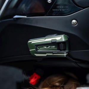 May include: A matte black motorcycle helmet featuring a dark tinted visor and a mounted olive green communication device on the side. The device displays the word JUKE in white lettering, accompanied by a black control dial and white graphic accents.