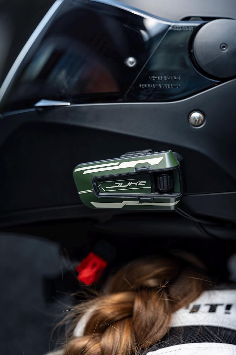 May include: A matte black motorcycle helmet featuring a dark tinted visor and a mounted olive green communication device on the side. The device displays the word JUKE in white lettering, accompanied by a black control dial and white graphic accents.