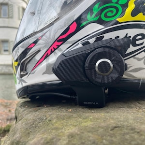 Może przedstawiać: Biały kask motocyklowy z czarno-ż&oacute;łto-r&oacute;żowym wzorem graficznym oraz zamontowanym z boku urządzeniem komunikacyjnym Sena o fakturze wł&oacute;kna węglowego. Kask posiada przezroczysty wizjer, a po prawej stronie widnieje napis Sixty.