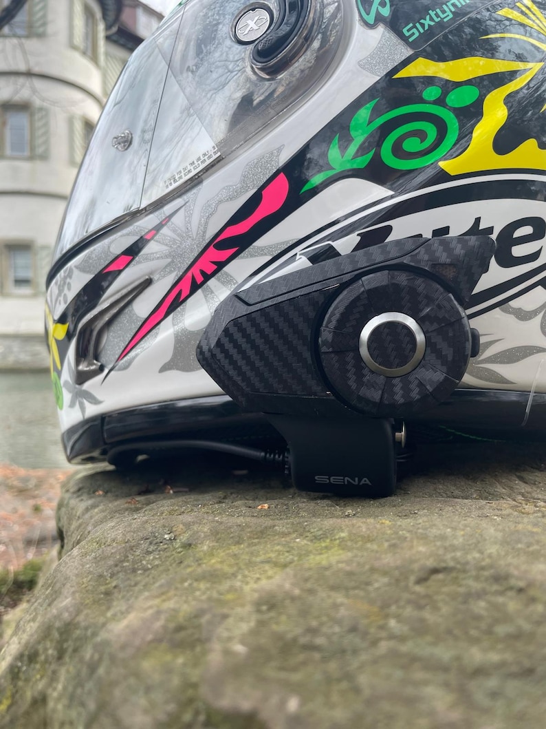 Może przedstawiać: Biały kask motocyklowy z czarno-ż&oacute;łto-r&oacute;żowym wzorem graficznym oraz zamontowanym z boku urządzeniem komunikacyjnym Sena o fakturze wł&oacute;kna węglowego. Kask posiada przezroczysty wizjer, a po prawej stronie widnieje napis Sixty.