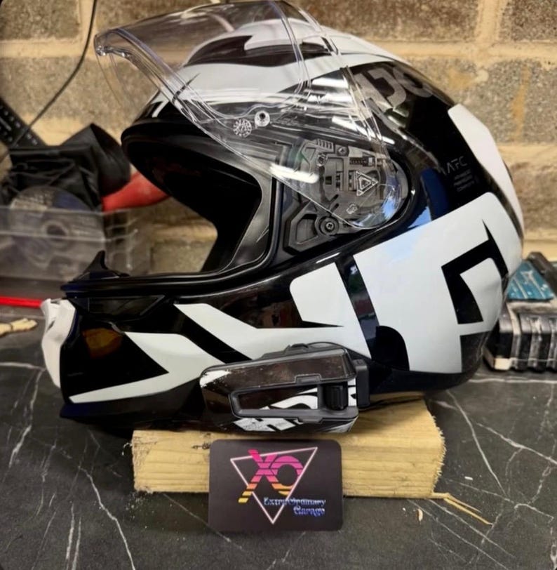 May include: A black and white full-face motorcycle helmet with a clear visor and a mounted communication device on the side. A small black rectangular sign with a pink and yellow geometric logo and the text Extraordinary Garage is placed in front.