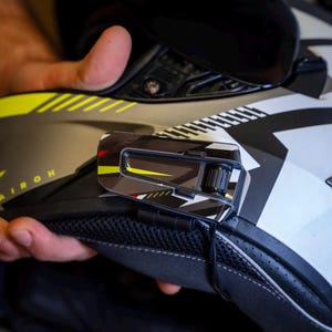 May include: A motorcycle helmet with a communication device attached. The helmet is primarily black, white, and gray, with yellow accents. The communication device is black and silver with red and yellow details.