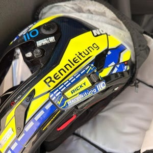 May include: A yellow, blue, and black racing helmet featuring the text Rennleitung, Ricky, and 110. The helmet has a dark visor, carbon fiber detailing, and a prominent yellow side panel with blue accents and white dashed lines.